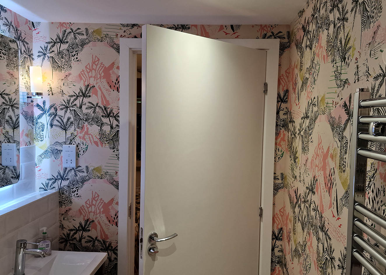 Milton & King Queen Palm wallpaper installed in a bathroom. Green palm trees with pink coloured shapes on white background.