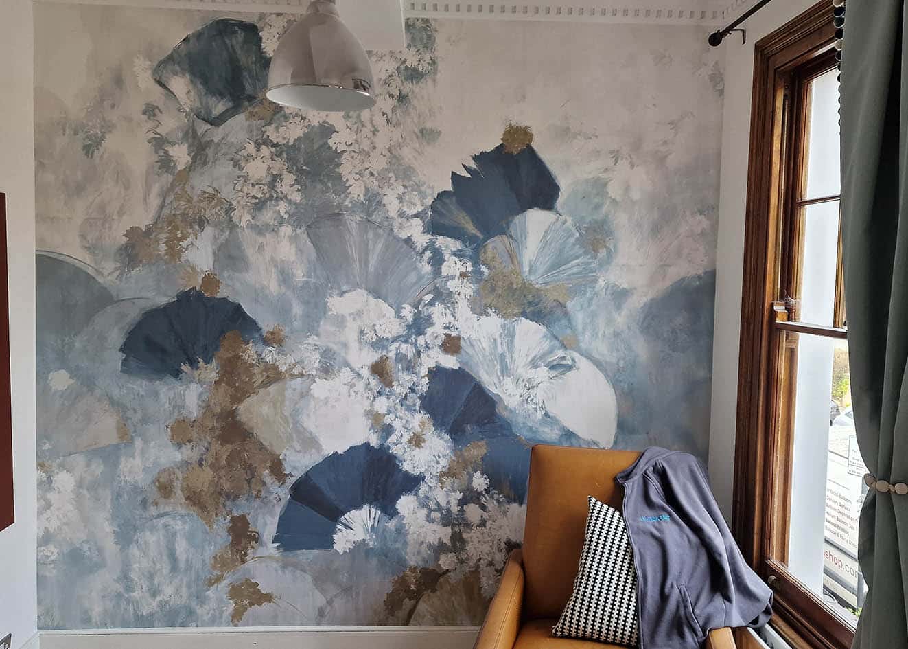 Iizzo wall mural installation. The wall mural is in shades of blue and golden with a large abstract flowers, It is next to a window in a room and there is n armchair in yellow with a Bluespec Decorating Limited logo jacket on it.