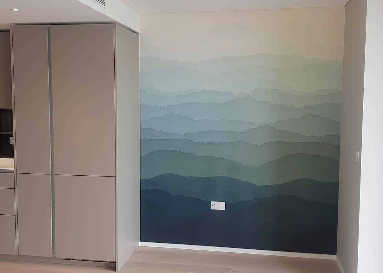 Wrey by Zoffany wall panel installed by our wallpaper hangers in a open plan living room. The wall panel is has overlayed layers in shades of blue with ombre effect. There is a kitchen unit on left.
