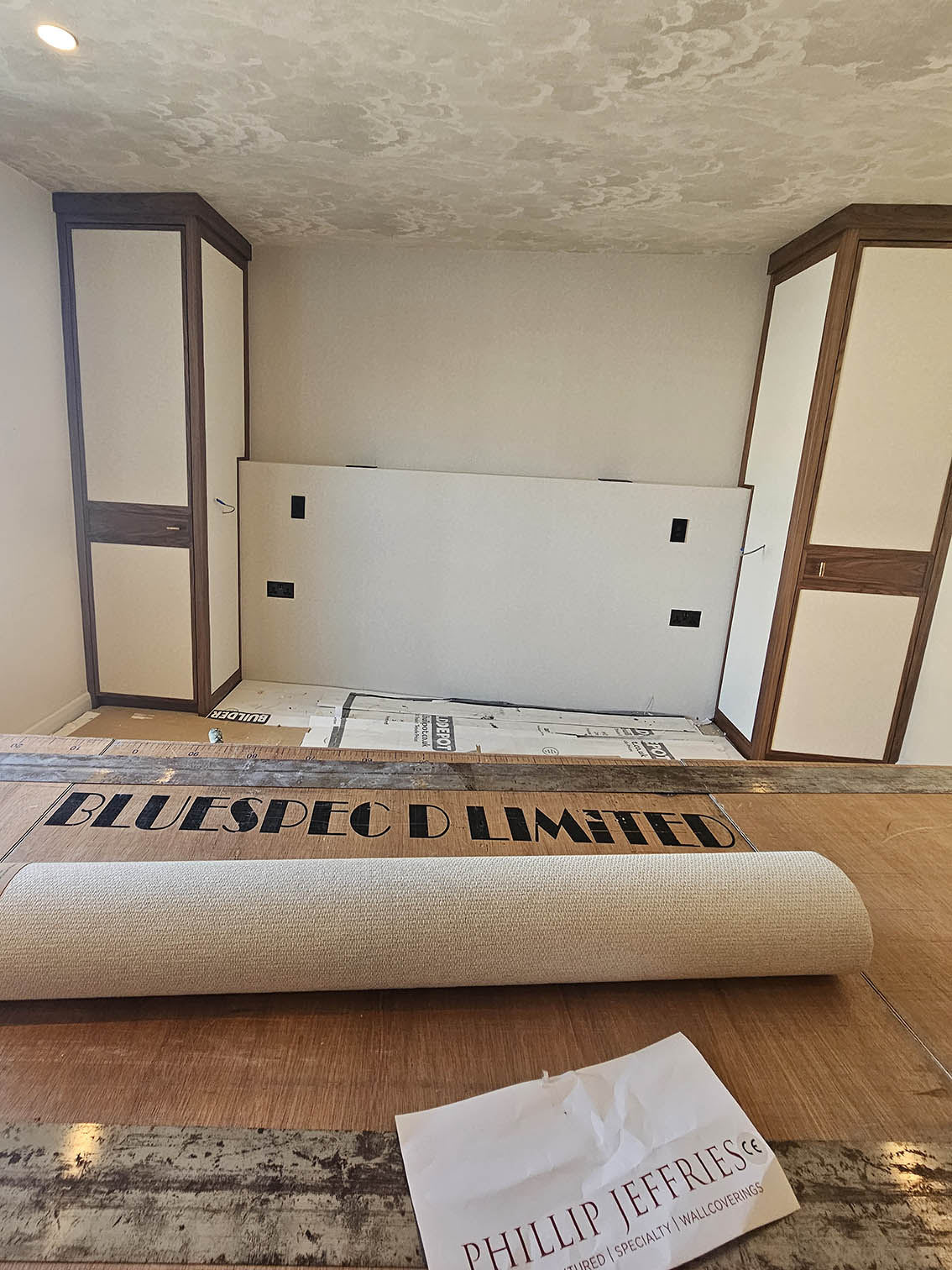 bluespec-decorating-waork in progress Wallpapered cabinet doors. A wallpapering tablewith a sign Bluespec Limited. Wallpaper roll and a Phillip Jeffries wallpaper instructions on it. The ceiling is wallpapered with clouds wallpaper.